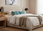 Teal Ari pillows on a bed with striped bedding and a grey throw