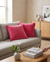 Two pink fringed velvet cushions on a grey sofa, a hand placing a book on the table