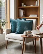 Teal fringed velvet cushions on a cream armchair beside a bookshelf and side table
