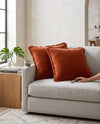 Two rust fringed velvet cushions on a grey sofa, a hand reaching for one
