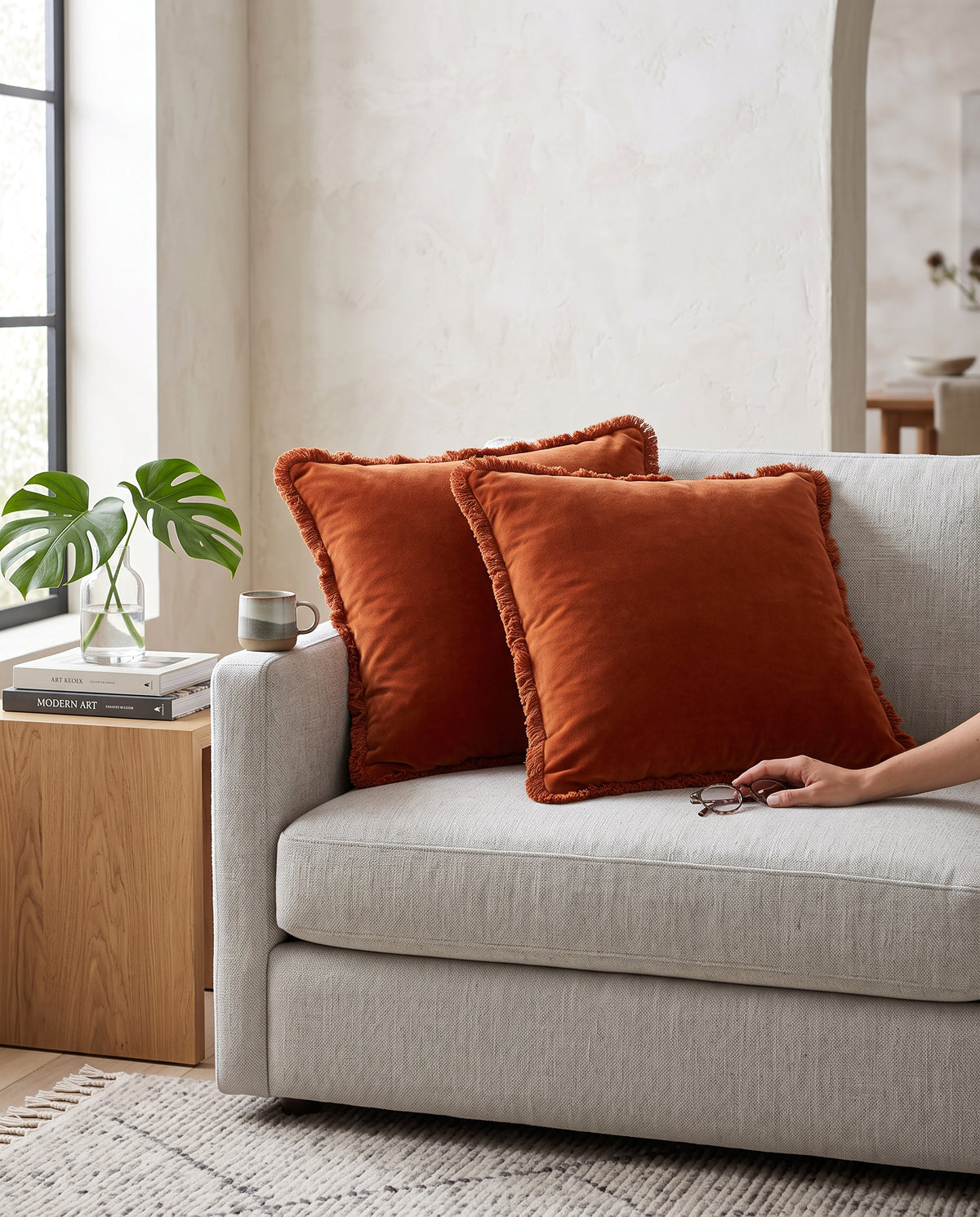 Two rust fringed velvet cushions on a grey sofa, a hand reaching for one