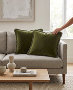 Two olive green fringed velvet cushions on a grey sofa, a hand adjusting one
