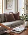 Dusty pink fringed velvet cushions on a grey sofa, a hand leafing through a magazine