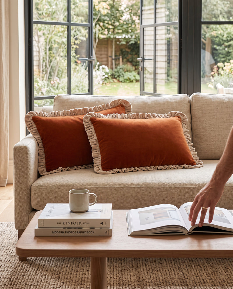Rust frill velvet lumbar cushions on a beige sofa, a hand turning pages of a book on the table