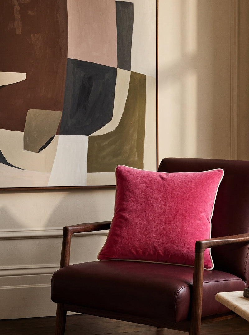 Pink piped velvet cushion on a burgundy leather chair in front of an abstract artwork