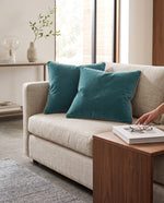Two teal piped velvet cushions on a cream sofa beside a wooden coffee table