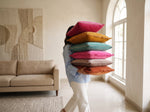 Person carrying a tall stack of colourful fringed velvet cushions through a bright living room