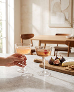Coffee coupe glasses with drinks on a counter beside figs and cheese, a hand lifting one