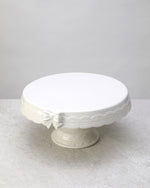 White glazed ceramic cake stand with a lace-textured edge and bow detail, on white