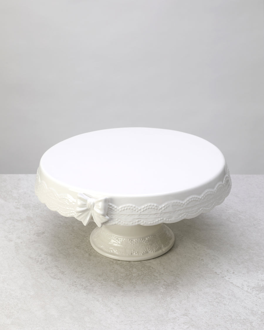 White glazed ceramic cake stand with a lace-textured edge and bow detail, on white