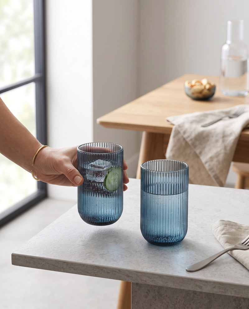 Blue ribbed highball glasses on a counter, a hand picking up one with cucumber water