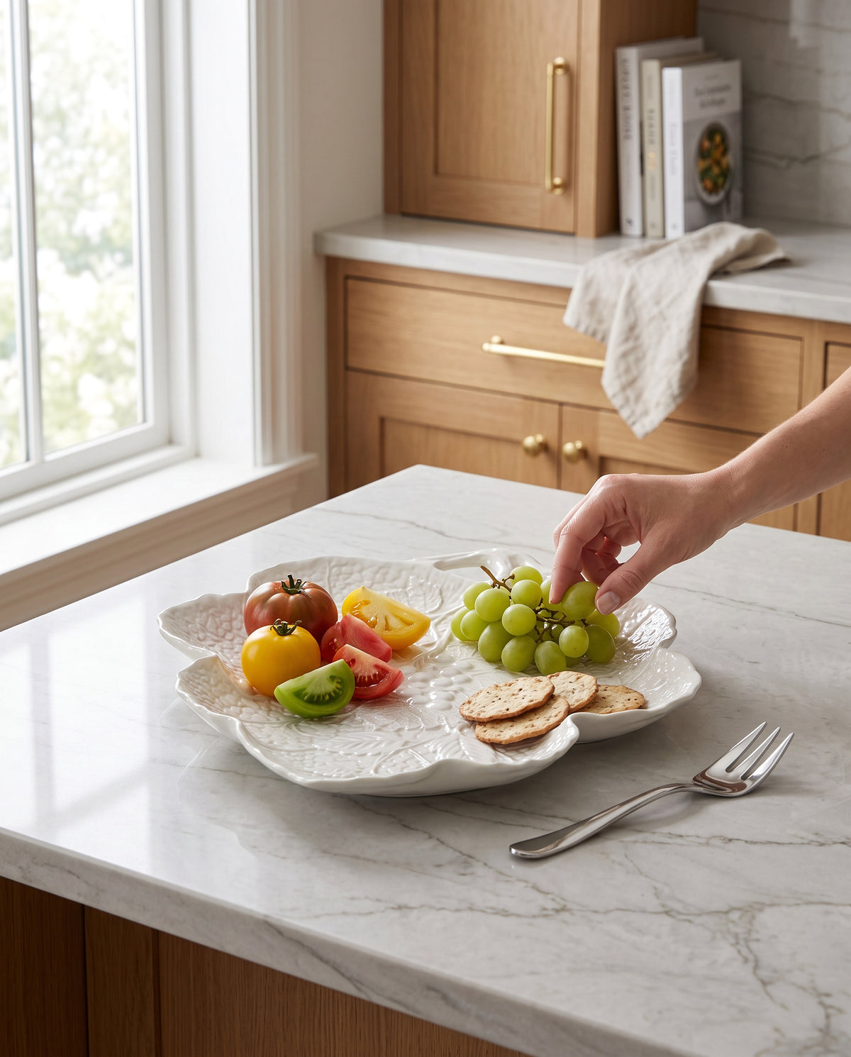 White porcelain leaf plate with fruit and cheese on a kitchen counter, a hand taking a grape