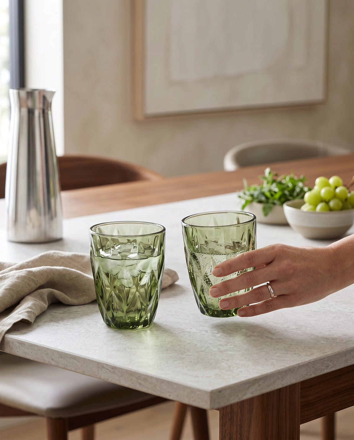 Olive diamond cut tumblers on a dining table beside a bowl of grapes, a hand holding one