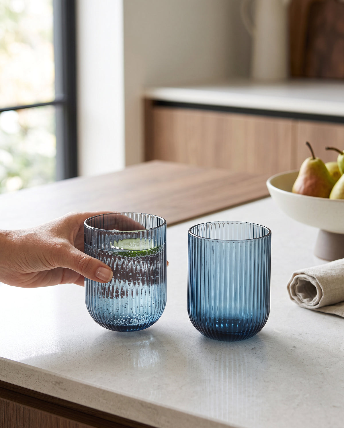 Blue ribbed tumblers on a kitchen counter beside a bowl of pears, a hand lifting one