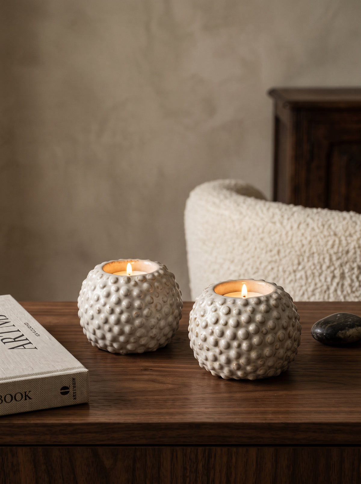 Two textured white ceramic tea light holders glowing on a wooden side table beside books