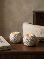 Two textured white ceramic tea light holders glowing on a wooden side table beside books
