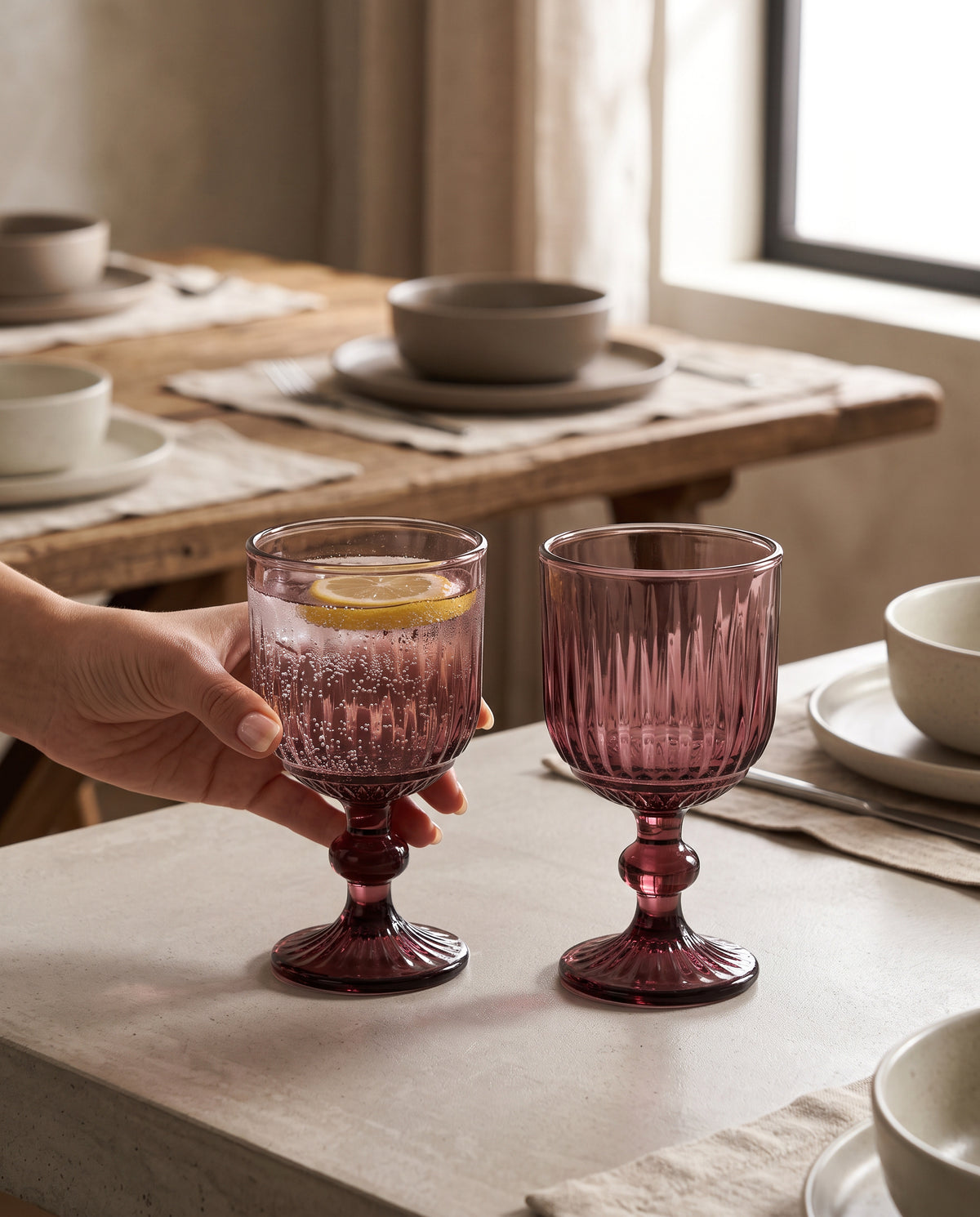 Burgundy ribbed wine glasses on a set table, a hand lifting one with a lemon slice