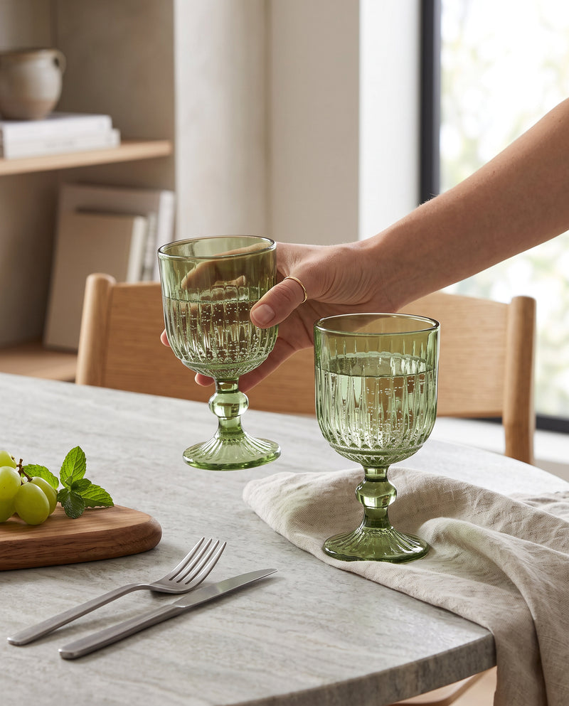 Hand pouring wine into green ribbed cut wine glasses at a table
