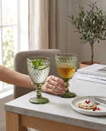 Olive green diamond wine glasses with drinks on a side table, a hand holding one