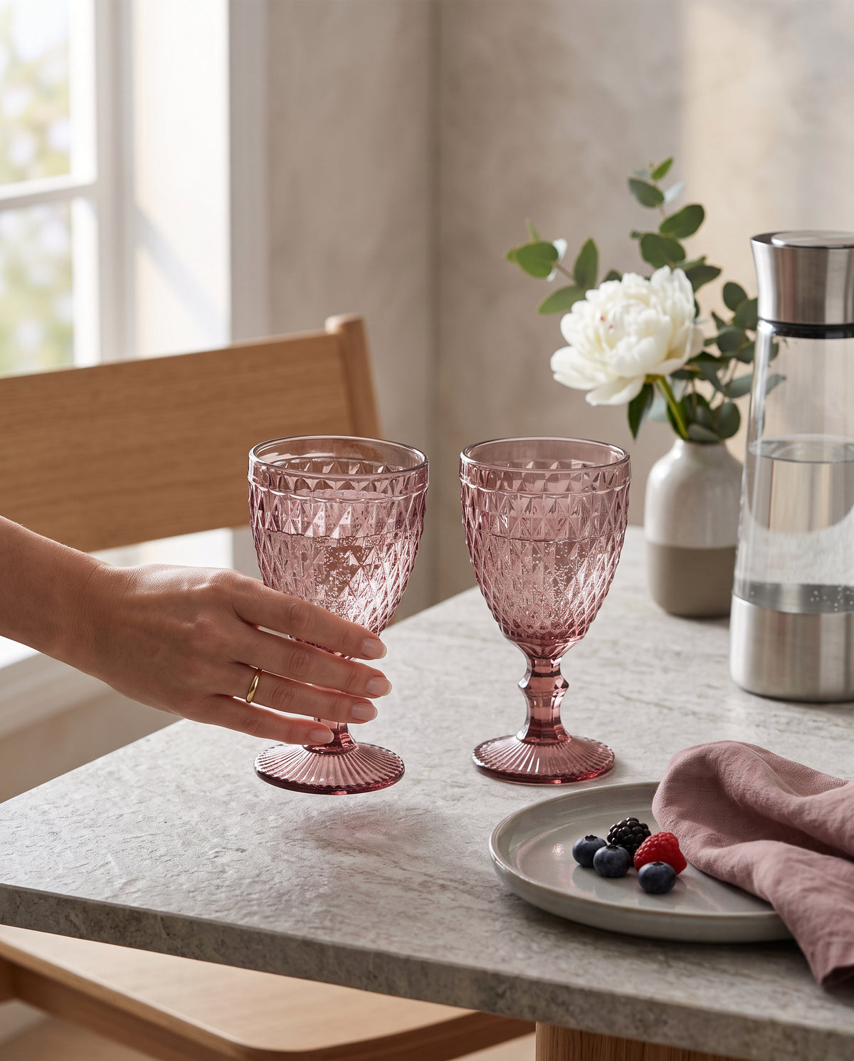 Pink diamond wine glasses on a table with berries and flowers, a hand holding one