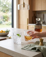 Hand holding a whiskey glass on a kitchen counter beside the decanter