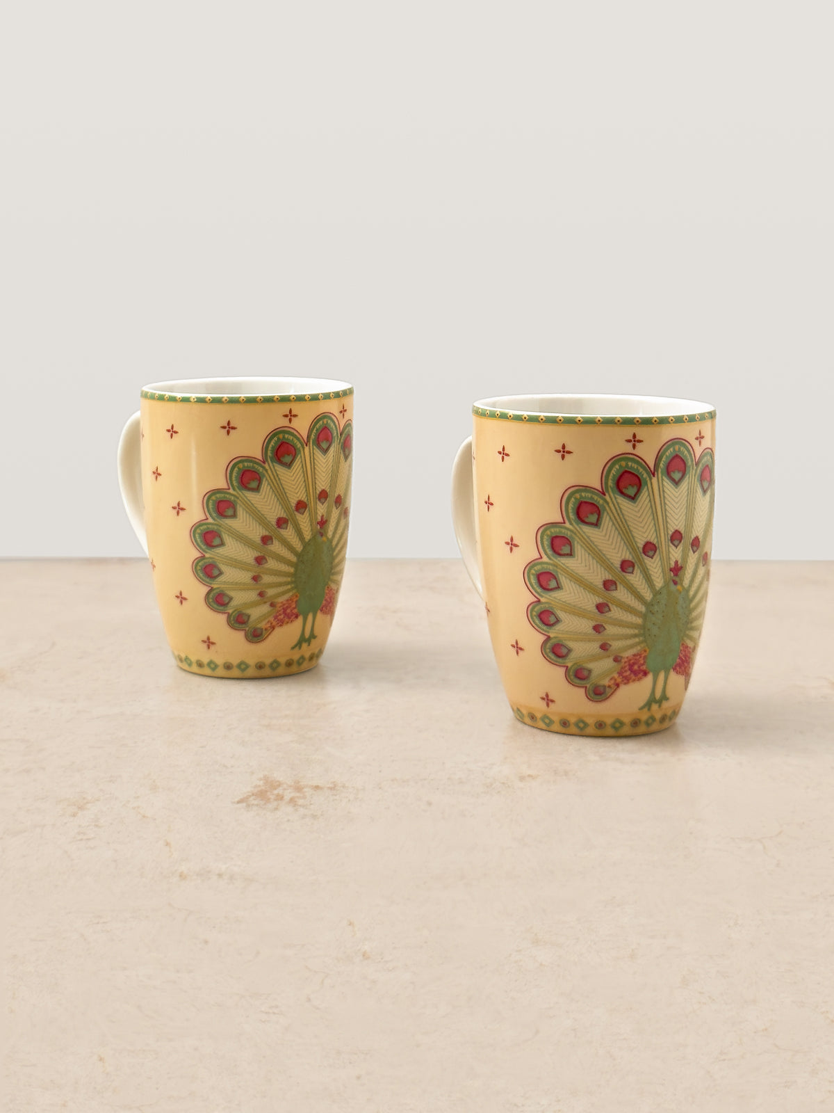 Two tall Mor Bagh porcelain coffee mugs in Dawn with a peacock feather print, front view