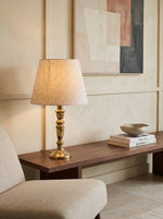 Slender gilded column lamp with a linen shade glowing on a wooden console