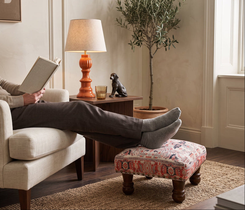 Red print square foot stool under resting feet in a reading corner beside a lamp