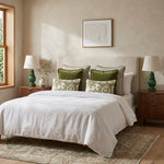 Olive green fringed velvet cushions on a white bed between green bedside lamps