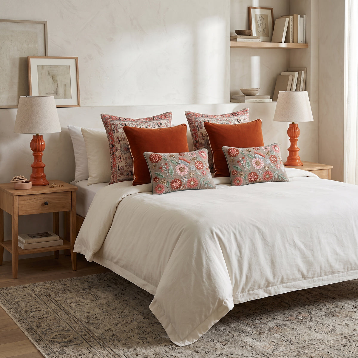 Beige velvet Dori pillows with rust cushions on a white bed between coral lamps